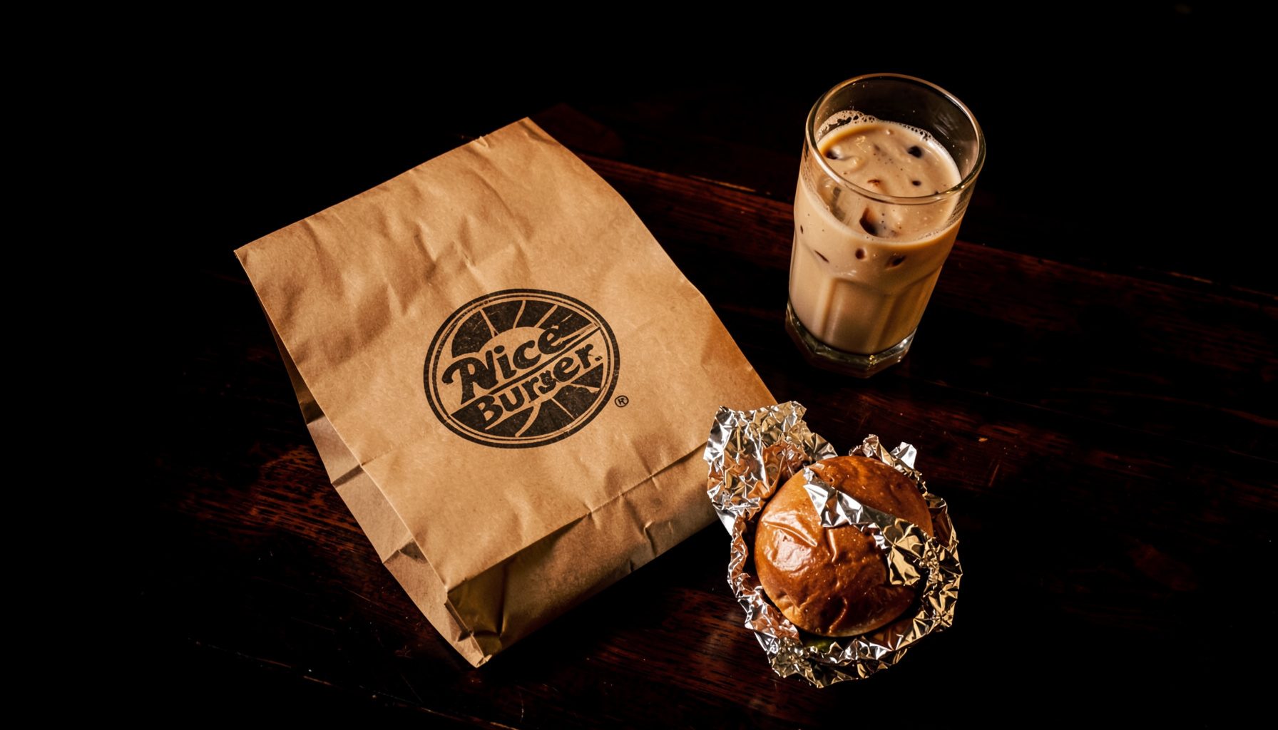 A warm overhead shot of a brown paper delivery bag with the Nice Burger logo stamp, sitting on a dark wooden table beside fries and a shake